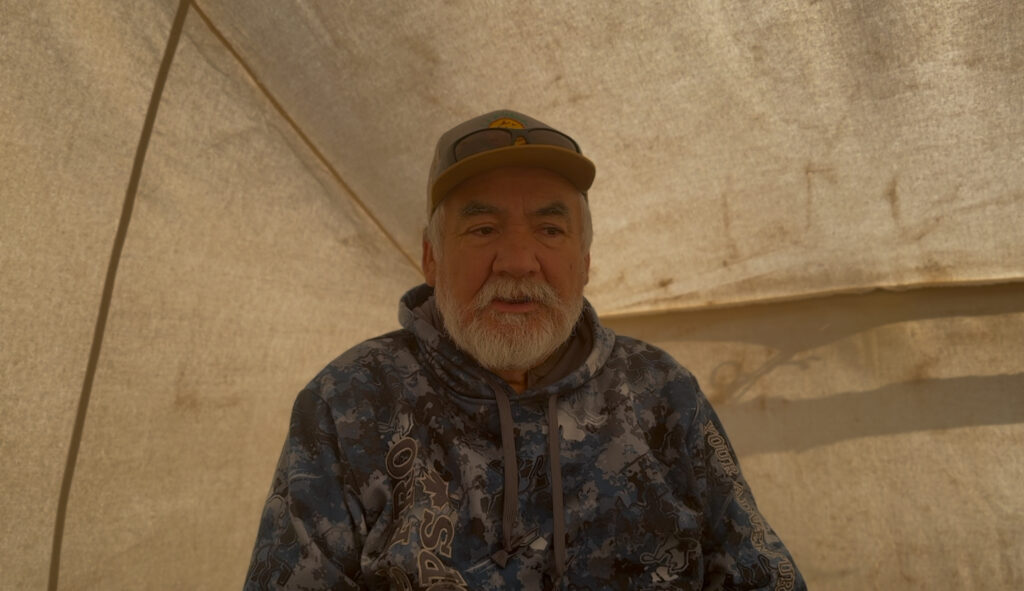 A man with a white beard and a calm expression sits inside a softly lit canvas tent. He wears a camo-patterned hoodie and a hat with sunglasses perched on top, creating a casual and reflective atmosphere. This moment captures an interview with Johnny Saganash, focusing on his thoughts about the Walking Out ceremony.