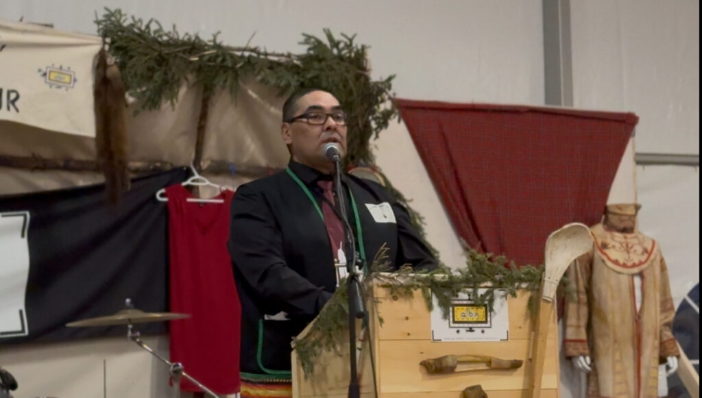 A man stands at a podium decorated with greenery and wooden elements, delivering an emotional farewell speech. He wears a formal outfit, and his expression reflects the gravity of the moment. Behind him are cultural symbols, including a traditional outfit and red fabric. His words resonate deeply, highlighting the wisdom gained from the elders' gathering and the comfort it brings in embracing aging.