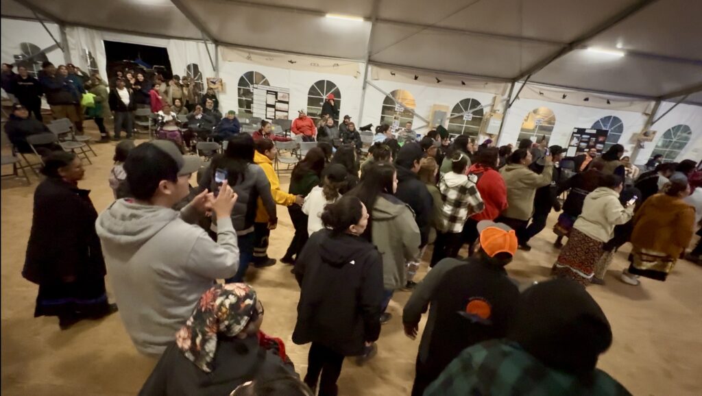 A large group of people fills the dance floor inside a tent, celebrating together. The crowd includes a mix of people, some taking photos and videos while others join in the dance. The energy in the room is lively, with everyone letting go of their shyness and fully participating in the celebration.