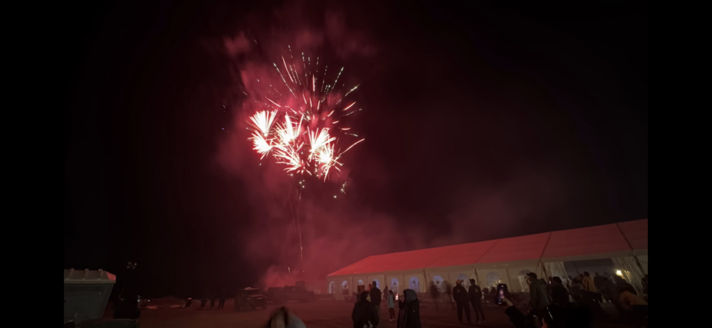 A vibrant fireworks display lights up the night sky with bursts of red and white, as people stand near a large tent, watching and capturing the moment on their phones. The scene is filled with excitement, marking the grand finale of the event.