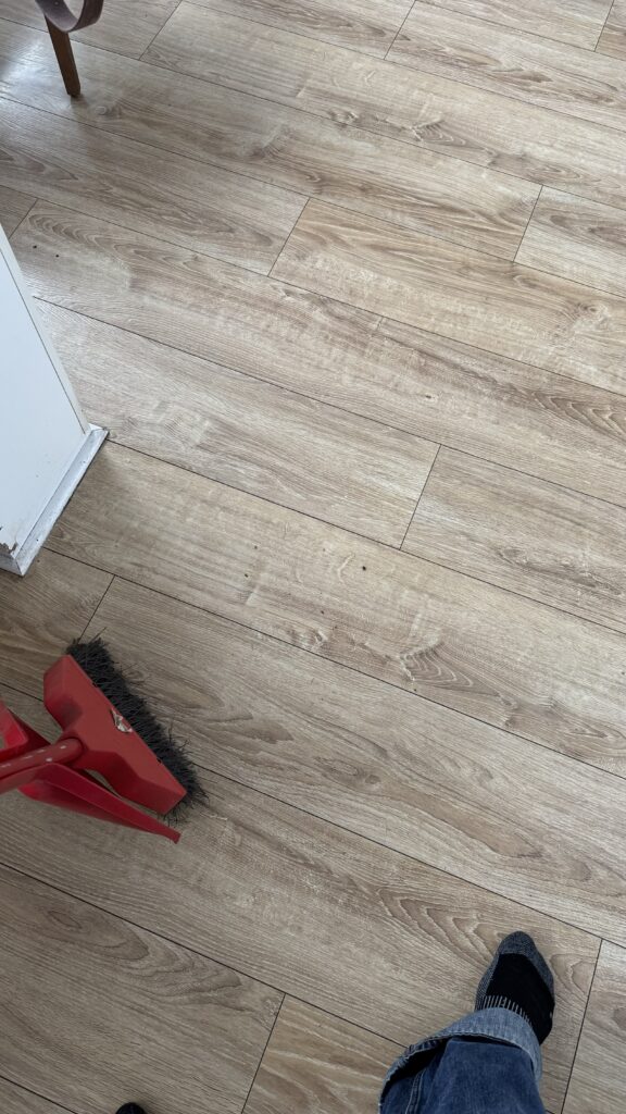 A light wood-colored laminate floor with a person’s foot in a black sock and jeans visible at the bottom right. In the bottom left, a red broom and dustpan are shown, as if mid-cleaning. The room appears clean with minimal dust or debris on the floor.