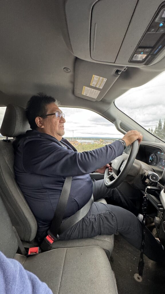 A man wearing glasses and a dark sweatshirt is seated in the driver’s seat of a vehicle, holding the steering wheel with both hands. He is buckled in with a seatbelt, and the view outside the window shows a cloudy sky and trees in the distance. The interior of the car, including the dashboard and roof, is visible.