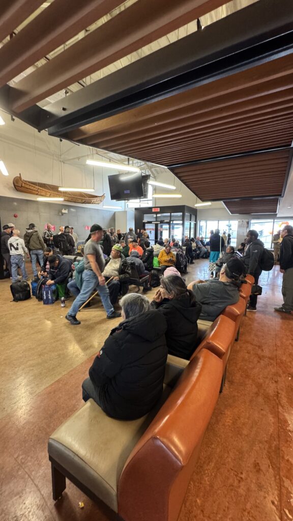 A crowded airport waiting area filled with people, many sitting on chairs or benches while others stand or walk through the space. Most people appear bundled in coats, and luggage is scattered around them. The ceiling features wooden beams, and a traditional canoe is mounted on the wall. The scene captures the busy atmosphere after an event, with people preparing to travel.