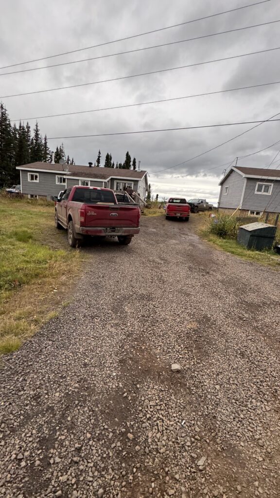 A gravel driveway leading up to a small gray house with trucks parked along the sides. Another gray house is visible on the right side, and a few trees stand in the background beneath a cloudy sky. The scene appears to be in a rural area, with signs of community gathering evident from the parked vehicles.