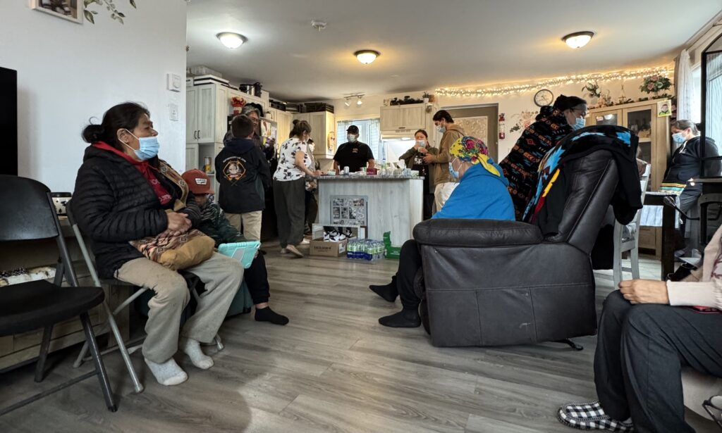 A group of people gathered inside a home, many wearing face masks. Some are seated on chairs and recliners, while others stand near a kitchen island in the background. The room is decorated with string lights and a mix of family photos and floral arrangements. A few people are passing out masks, while others engage in conversation, creating a warm and communal atmosphere.