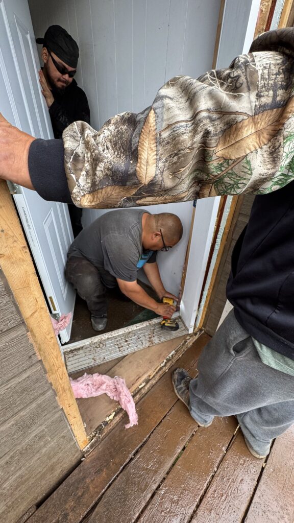 A man kneels at a doorway, using a drill to remove the door frame. Another man stands in the background, watching as the work is being done. The door frame has been partially dismantled, with pieces of pink insulation visible on the wooden deck. The scene shows preparations for accommodating pallbearers carrying a casket.