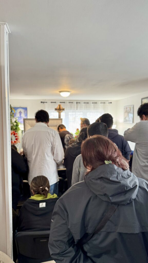 A crowded room filled with people standing and sitting, as they pay their respects during a funeral service. A casket is visible at the front of the room, with mourners gathered around it. A cross hangs on the wall in the background, and the room is simply decorated. The atmosphere is somber as visitors move through the space.