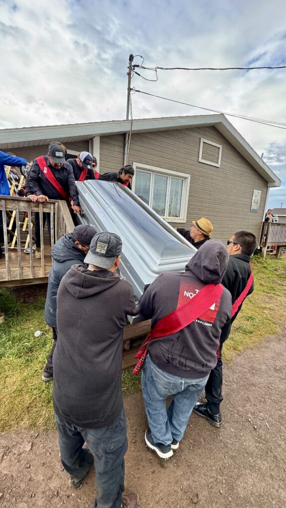 A group of pallbearers, wearing black hoodies and red sashes, carefully carry a casket down the steps of a house toward a waiting hearse. The casket is silver, and the men work together to lower it from the porch. The house is a modest home with brown siding, and the sky is partly cloudy.
