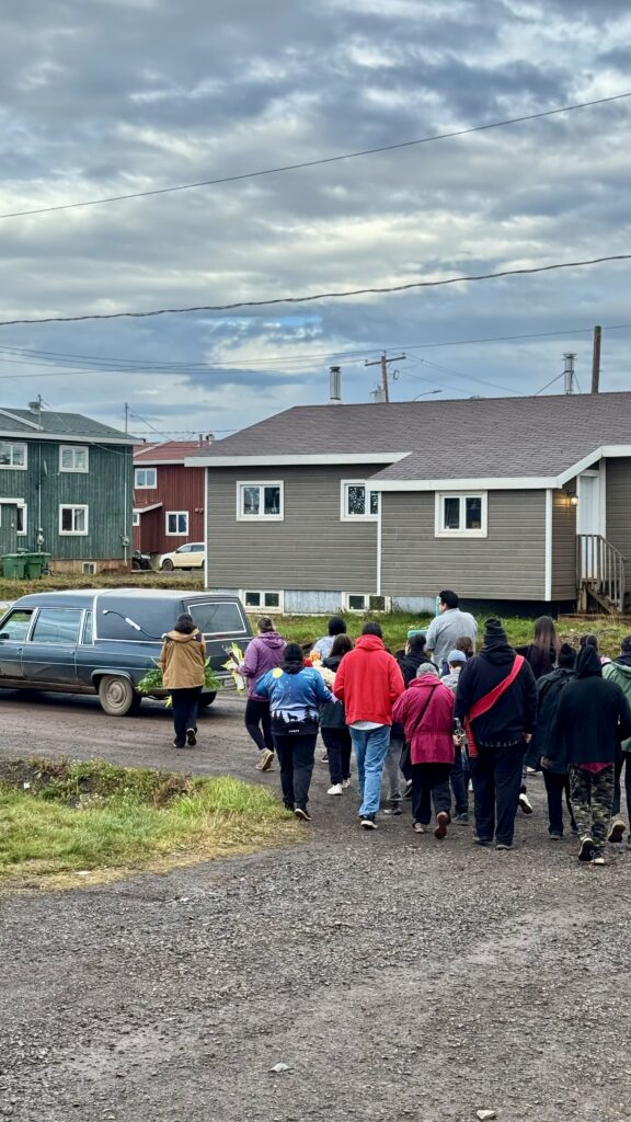 A funeral procession follows a hearse down a dirt road, with people walking closely behind it. Some carry flowers, and others wear red sashes over their clothing. The scene takes place in a residential area with modest houses and overcast skies, reflecting the somber mood of the procession.