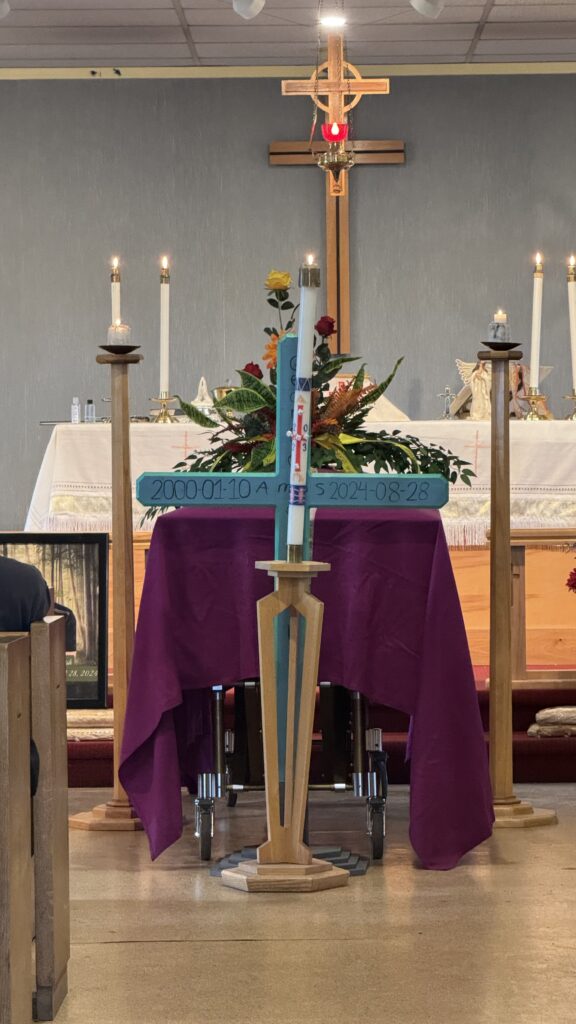 The interior of a church with a casket draped in a purple cloth at the front, adorned with a large bouquet of flowers. In front of the casket, a wooden cross with dates "2000-01-10" and "2024-08-28" is displayed. Lit candles and religious symbols, including a crucifix on the wall, surround the altar, creating a solemn and reverent atmosphere.