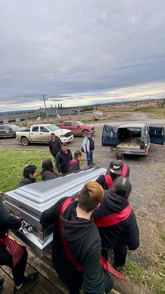  A group of pallbearers, wearing black with red sashes, carry a silver casket down the steps of a church and towards an open hearse. The scene takes place in a small community, with several trucks parked nearby and houses visible in the background under a cloudy sky. The mood is solemn as the service concludes and the casket is prepared for transport.