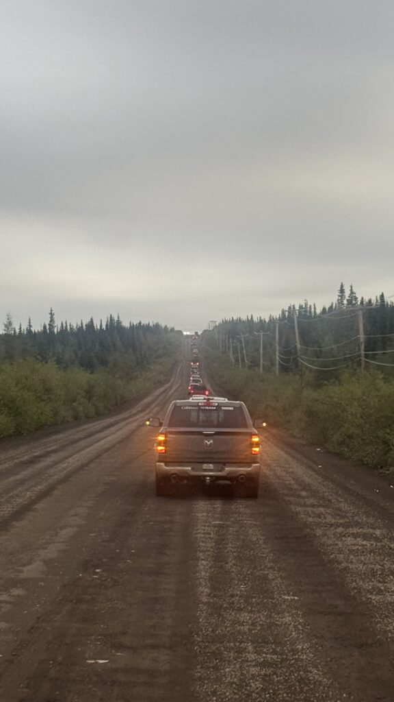 A long line of vehicles drives down a dirt road, forming a procession as they make their way through a forested area. The cars have their lights on, and the road stretches into the distance, surrounded by trees and utility poles. The procession is part of a traditional funeral, passing through the village before heading to the cemetery.