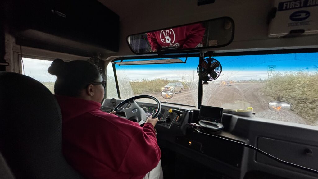 The view from inside a vehicle, showing a woman in a red sweatshirt driving a bus along a dirt road. Vehicles traveling in the opposite direction have pulled over, stopping to allow the funeral procession to pass. The windshield is covered with dust, and the scene outside shows a cloudy sky and a few vehicles on the road.