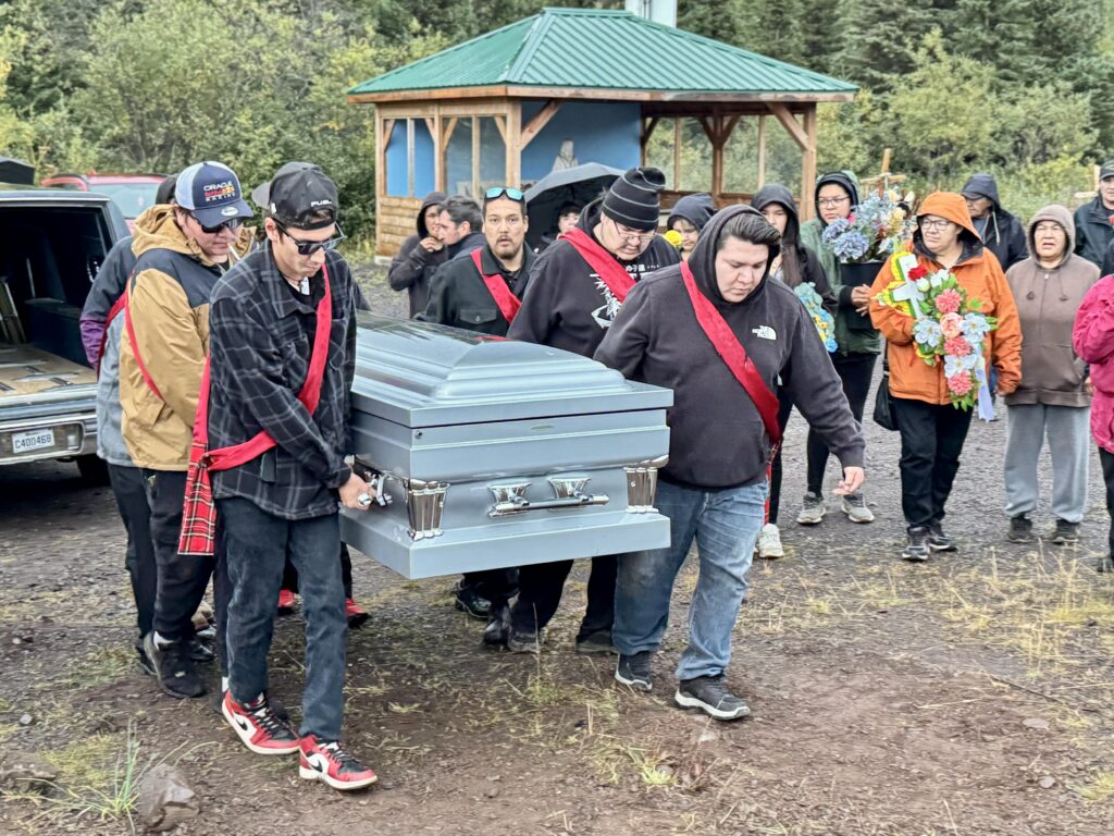 A group of pallbearers, wearing red sashes, carry a silver casket from the hearse at a cemetery. Family members follow closely behind, some holding floral wreaths. The scene takes place outdoors, with a small shelter in the background and a mix of emotions on the faces of the mourners.