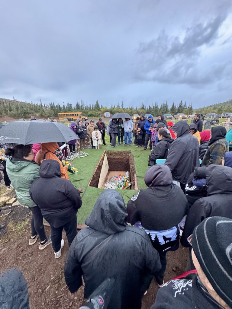  A large group of mourners gathers around an open grave at a cemetery, some holding umbrellas as light rain falls. The grave is lined with green turf, and floral arrangements can be seen inside the grave. People stand in silence, offering prayers and support to one another.