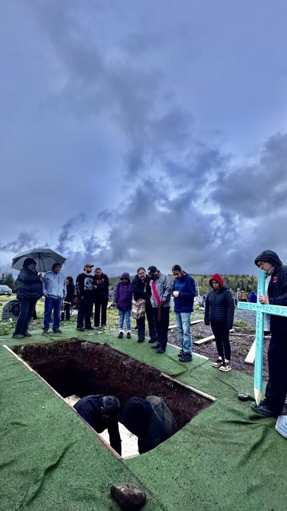 A group of mourners stands around an open grave at a cemetery, with some holding umbrellas as a light rain begins to fall. Two men inside the grave prepare to lower the casket, while a wooden cross marked with the name and dates of the deceased is held by one mourner. The sky is dark and overcast, reflecting the somber mood.