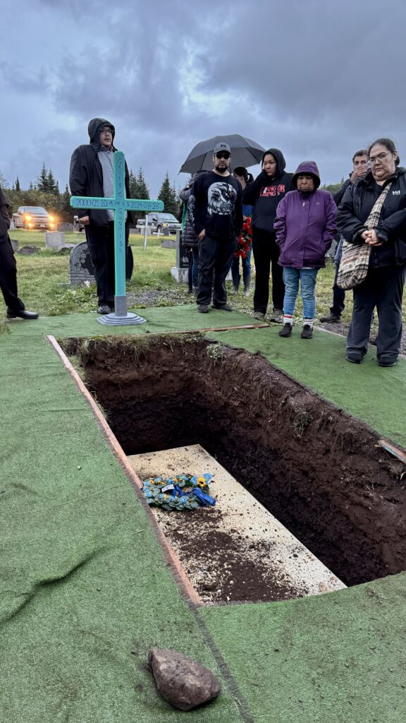 A group of mourners stands around an open grave, where a blue floral wreath rests on the casket at the bottom. One mourner holds a cross painted with the name and dates of the deceased. The sky is dark and overcast, and the mood is solemn as people reflect and offer comfort.
