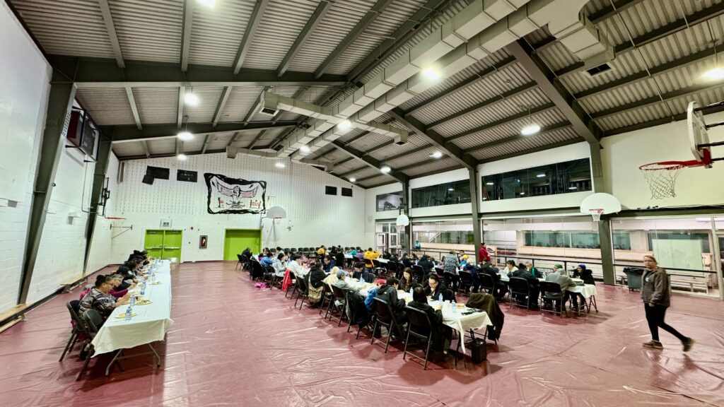 A large gathering in a gymnasium, where tables are set for a feast following the funeral ceremony. People are seated along long rows of tables, sharing a meal and conversations. The atmosphere is a mix of somber reflection and warmth as family and friends come together to remember the deceased. The gym's high ceilings and basketball hoops are visible, and a large banner hangs on the back wall.