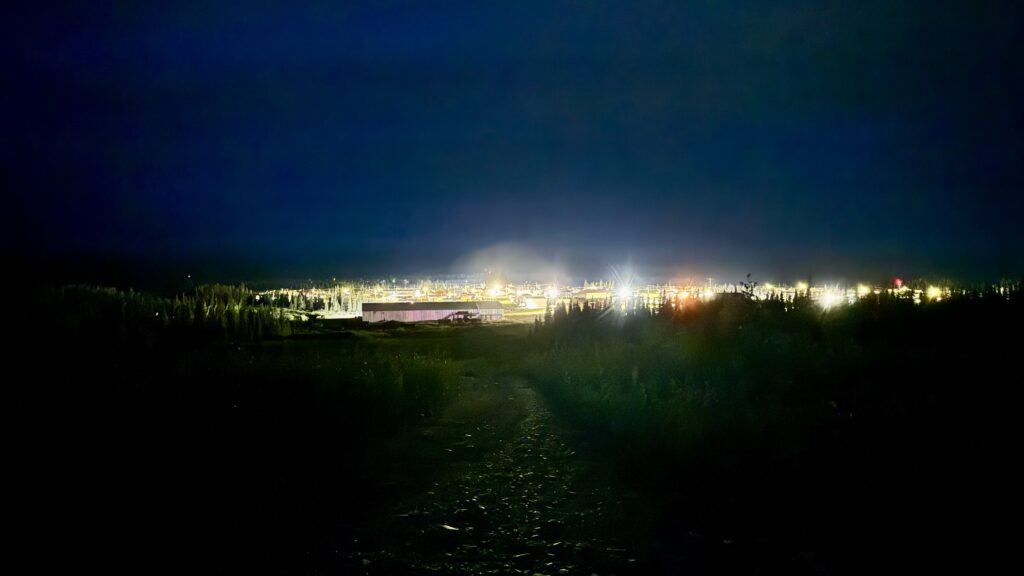  A nighttime view of a village illuminated by bright lights in the distance, seen from a hill surrounded by darkness. The sky is almost entirely black, while the lights below create a soft glow over the trees and buildings, evoking a sense of quiet reflection.