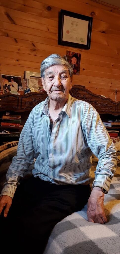 An elderly man, identified as Matthew Mameamskum, sits on a bed in a warmly lit room with wood-paneled walls. He is wearing a light striped button-up shirt and looking directly at the camera with a calm expression. Behind him are family photos and books on a wooden dresser, adding a personal and cozy atmosphere to the scene.