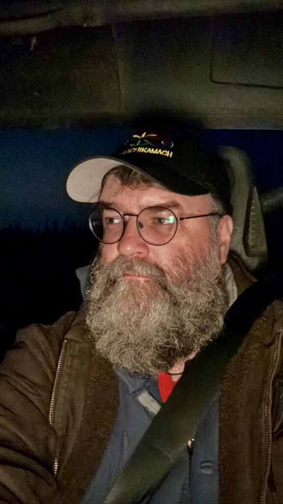 A bearded man, wearing glasses and a cap with the word "Kawawachikamach" embroidered on it, sits in a vehicle at night. He is looking ahead with a thoughtful expression, illuminated by the dim light inside the vehicle. A seatbelt is visible across his chest, and the background outside is dark.