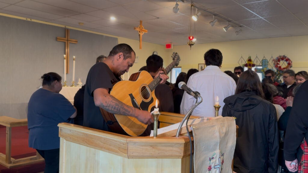 A man with a guitar, identified as Uapush, stands at the front of a church near the pulpit, playing a song during a funeral service. The congregation is gathered closely around, with many people standing in the pews. A lit candle sits on the pulpit, and a wooden cross is visible on the wall behind the altar. The emotional moment brings a sense of solemnity to the scene.
