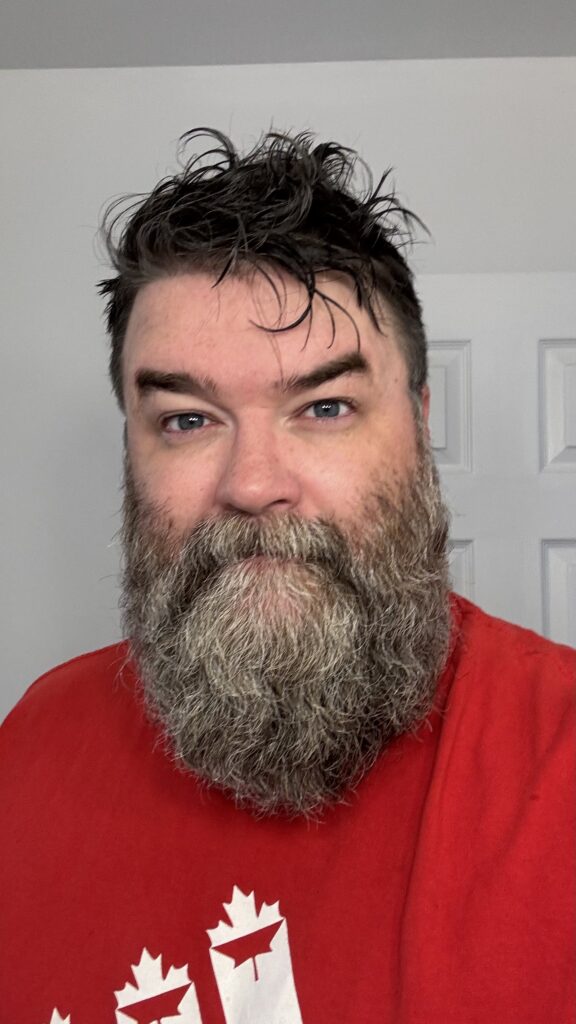 A man with a full beard and slightly wet, tousled hair takes a selfie after a shower. He is wearing a red shirt with a Canadian symbol on it, and he looks directly at the camera with a content expression. The background shows a plain white wall and door, suggesting the photo was taken at home while preparing for the day.