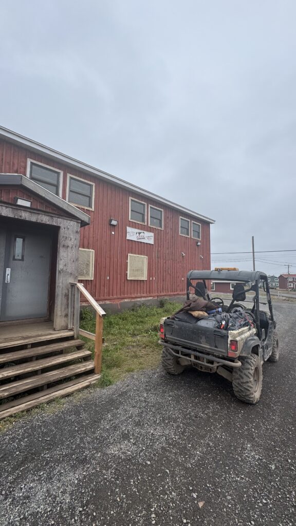 A red wooden building with the sign "Naskapi Development Corporation" is seen on a cloudy day. A rugged side-by-side vehicle is parked in front of the building, loaded with bags and gear. The building has boarded-up windows on the ground floor, and wooden steps lead up to the entrance. The vehicle, covered in mud, reflects its use in rough terrain.