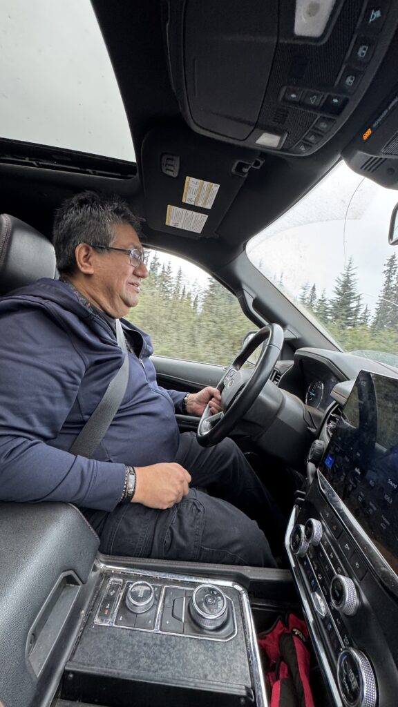 A man, Jimmy Shecanapish, is seen driving a large SUV, smiling and focused on the road. He is wearing a dark blue hoodie and glasses, with his seatbelt on. The interior of the vehicle is modern with a touchscreen display and controls on the console. Trees and the road are visible through the windows as they drive.