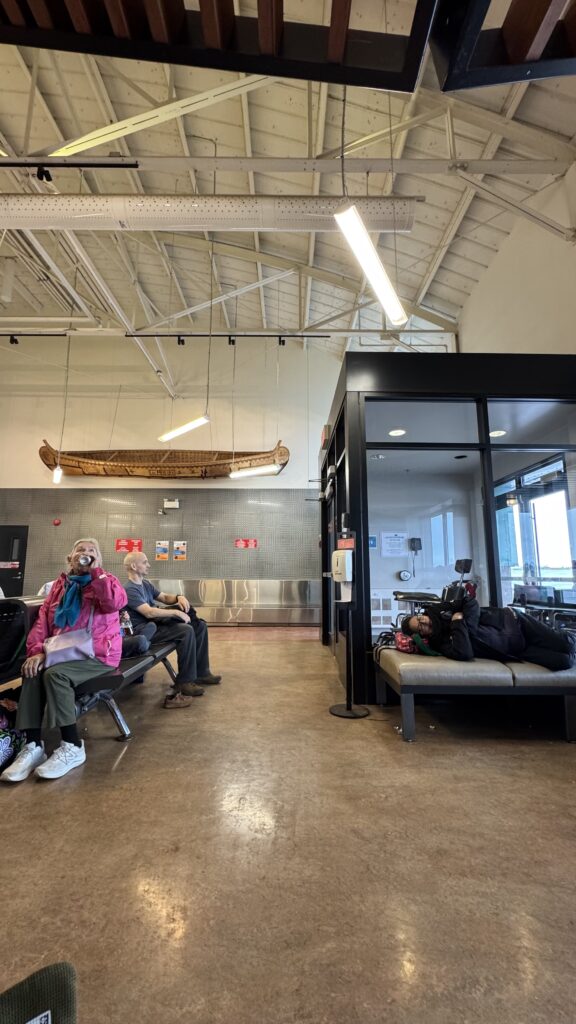 An airport waiting area with several people seated. A woman in a pink jacket drinks from a bottle, while a man in gray sits beside her. Another person is lying down on a bench in the foreground. Above the seating area hangs a traditional canoe, beautifully crafted and suspended from the ceiling. The canoe is a reminder of the unique cultural heritage of the community.