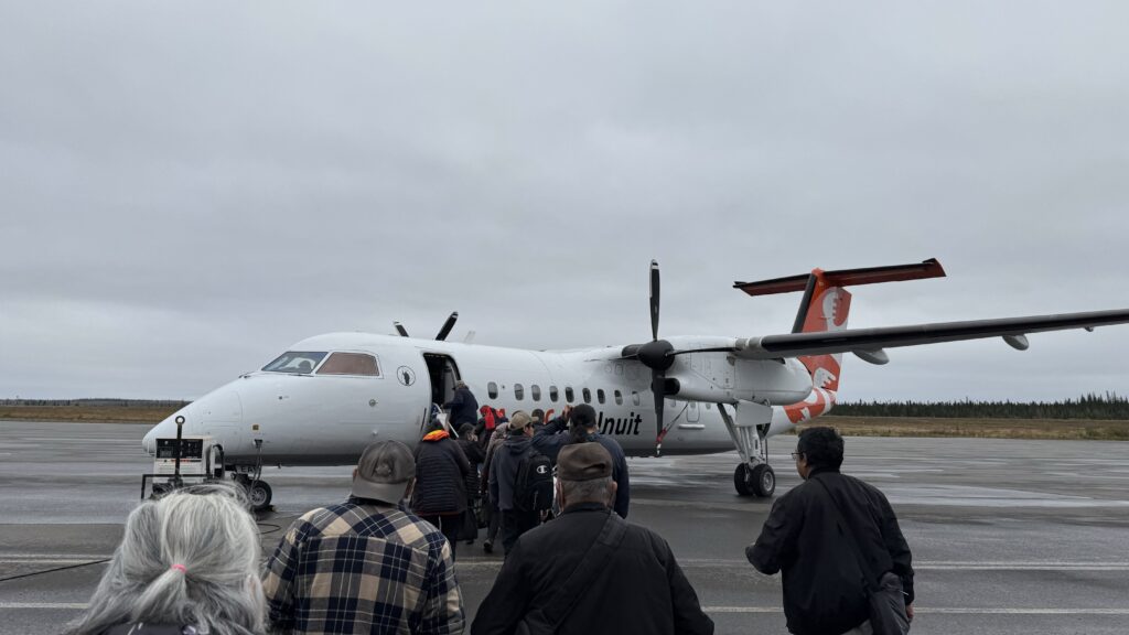 A group of people boarding a white Air Inuit plane on a cloudy day. The plane has two propellers, and the tail is painted with the Air Inuit logo in orange. The passengers are slowly walking in a line toward the aircraft, with a few people wearing hats and jackets, carrying small bags. The mood seems calm, but the overcast sky adds a somber feel to the moment.