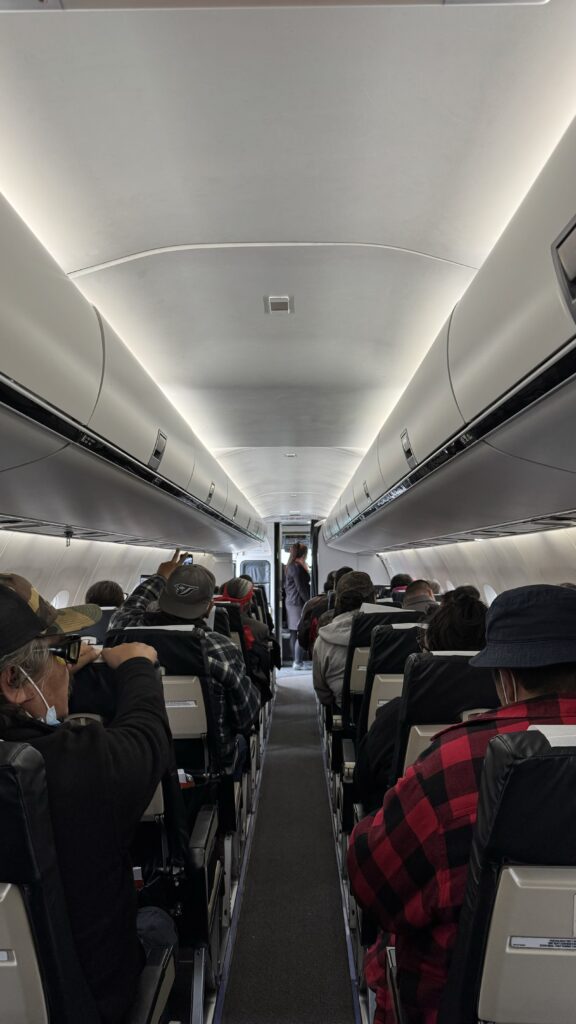 View from the back of a small airplane cabin, showing rows of passengers seated. Many are wearing hats, and one person is holding up a phone, perhaps to take a picture or video. The overhead compartments are closed, and the aisle leads to the front, where a flight attendant stands near the exit door. The mood feels quiet and reflective, with most passengers appearing calm.