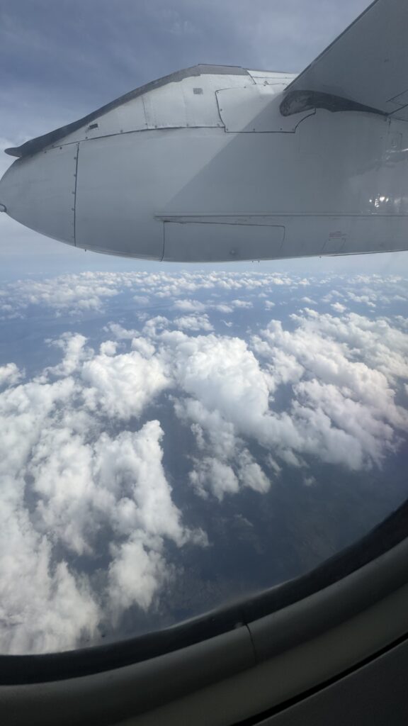 View from an airplane window showing part of the plane's wing and engine above a blanket of fluffy white clouds. The soft blue sky meets the horizon, and the clouds stretch out in various shapes and sizes. The scene evokes a sense of calm and peace, with the beauty of flying high above the earth.