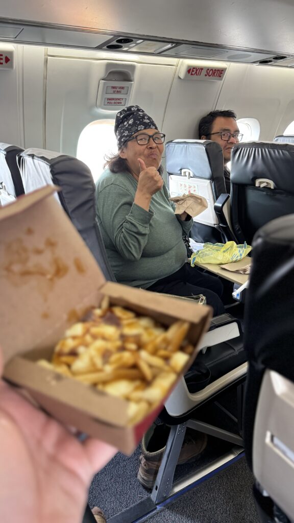 A person in a green sweater, sitting on a plane, smiling and giving a thumbs-up. In the foreground, a box of poutine is being held up, slightly blurred but clearly showing fries, cheese curds, and gravy. The person in the background seems to be enjoying the moment, creating a cheerful atmosphere. The plane's emergency exit signs are visible above the seat.