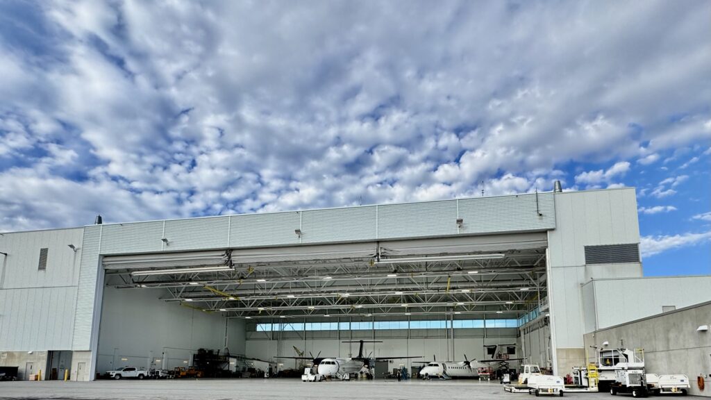 The exterior of a large airplane hangar with its massive doors open. Inside, several propeller planes are visible, along with ground support vehicles parked outside. The sky above is filled with scattered, fluffy clouds, creating a vibrant contrast with the industrial structure.