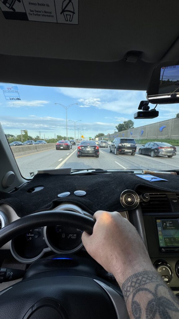 A view from the driver’s seat looking at a congested highway. The driver's hand, tattooed with a pattern, grips the steering wheel, while cars line up ahead in multiple lanes. The sky is blue with scattered clouds, and a GPS display shows the route to Quebec.