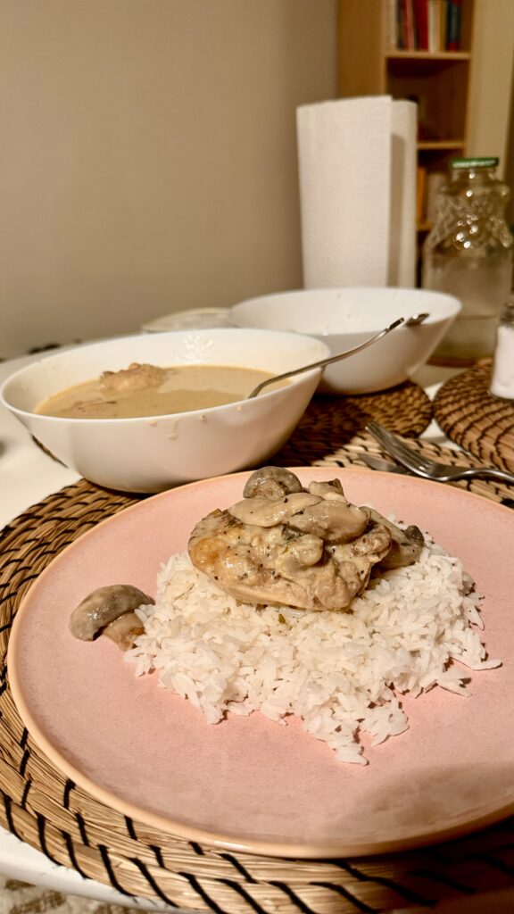 A plate of creamy mushroom chicken served over white rice on a pink plate. A bowl of the same dish, with a spoon resting inside, sits in the background on a woven placemat. The cozy dinner setup includes a paper towel roll and glass bottles nearby.