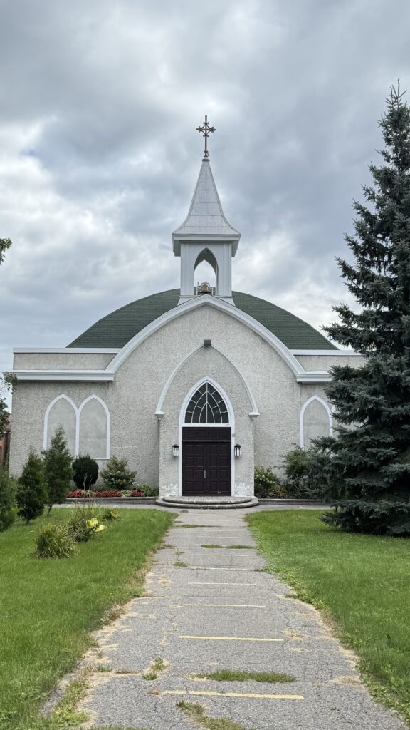 A small church with a domed green roof and a white steeple topped with a cross, seen from a straight-on view. The path leading to the dark wooden doors is cracked and weathered, flanked by green grass and small shrubs. The sky above is overcast, casting a soft light on the building.