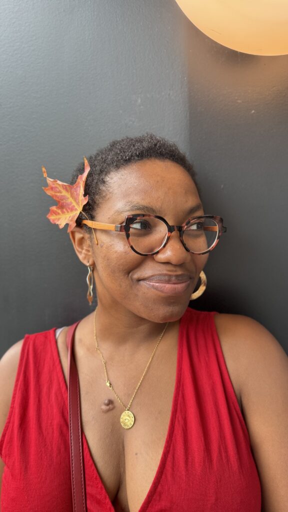 A woman with short hair smiles softly, wearing tortoiseshell glasses and a bright red sleeveless top. She has a fall leaf tucked behind her ear, and she’s accessorized with gold earrings and a pendant necklace. She’s standing against a dark background, with warm lighting illuminating part of the image.