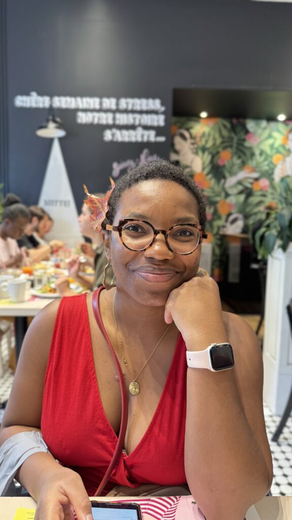 A woman with glasses and a red dress smiles softly while sitting in a restaurant. She has a fall leaf tucked behind her ear, and her hand rests against her chin. In the background, other people are dining, and a sign on the wall reads "Chère semaine de stress, notre histoire s'arrête."