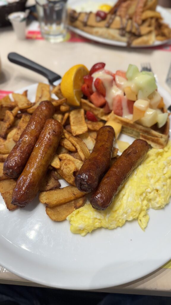 A hearty breakfast plate featuring scrambled eggs, golden brown breakfast potatoes, three sausages, and a fresh fruit-topped waffle. The waffle is garnished with strawberries, cantaloupe, and honeydew, drizzled with cream, and accompanied by a slice of lemon on the side.