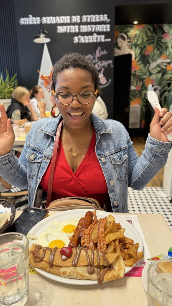 The same woman, now wearing a denim jacket over her red dress, sits in front of a hearty brunch. She is smiling and gesturing excitedly. Her plate includes sunny-side-up eggs, bacon, sausage, potato wedges, and a crêpe drizzled with chocolate and topped with a strawberry. The restaurant background features a sign with the phrase "Chère semaine de stress, notre histoire s'arrête."