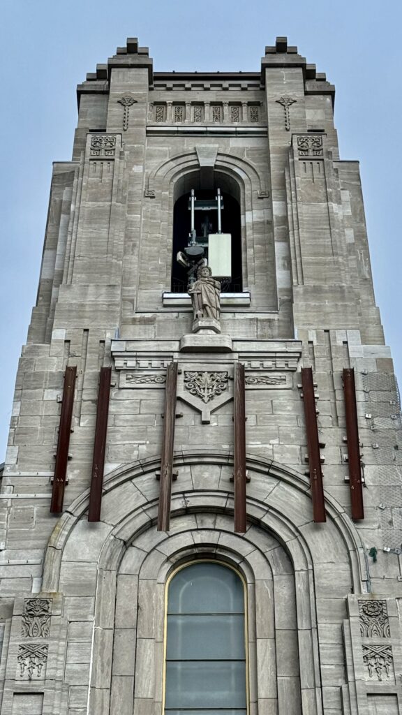 A tall stone church tower with a statue of a saint standing in a niche partway up. Above the statue is a large, modern cellphone transmitter, creating a striking contrast between the historical architecture and contemporary technology. The transmitter is prominent and unadorned, sitting visibly at the top of the tower.