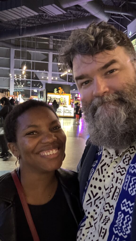 A smiling couple takes a selfie inside what appears to be a movie theater lobby. The woman is wearing a black leather jacket and the man has a thick beard and is wearing a patterned shirt. The background is lit with colorful lights and displays, likely from arcade games or movie posters. The atmosphere suggests they are about to enjoy or have just finished watching a movie.