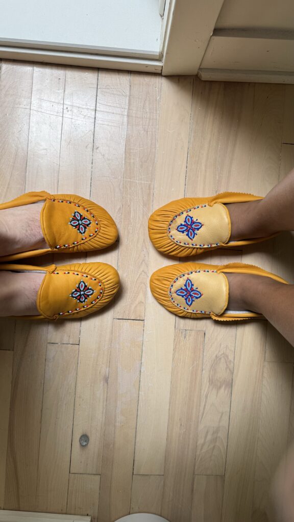 Two pairs of matching yellow moccasins with intricate beadwork on the top, featuring a blue, red, and white floral design. The moccasins are being worn by two people standing on a light wooden floor, their feet positioned towards each other. The moccasins are traditional Naskapi footwear, beautifully crafted.