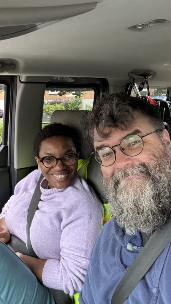 A smiling couple takes a selfie inside a car. The woman is seated in the passenger seat, wearing a light purple sweater and glasses, while the man is in the driver's seat, sporting a beard, glasses, and a blue hoodie. Both are wearing seatbelts, and the car's interior is visible with a bit of the outside background showing through the windows.