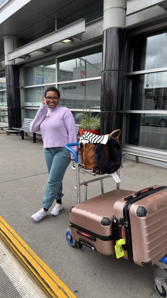  A woman stands outside an airport terminal, smiling and waving while posing beside a luggage cart. She is wearing a light purple sweater, teal pants, and white sneakers. The cart holds two rose-colored suitcases, a brown handbag, and a neck pillow. The airport's glass windows and modern design are visible in the background.