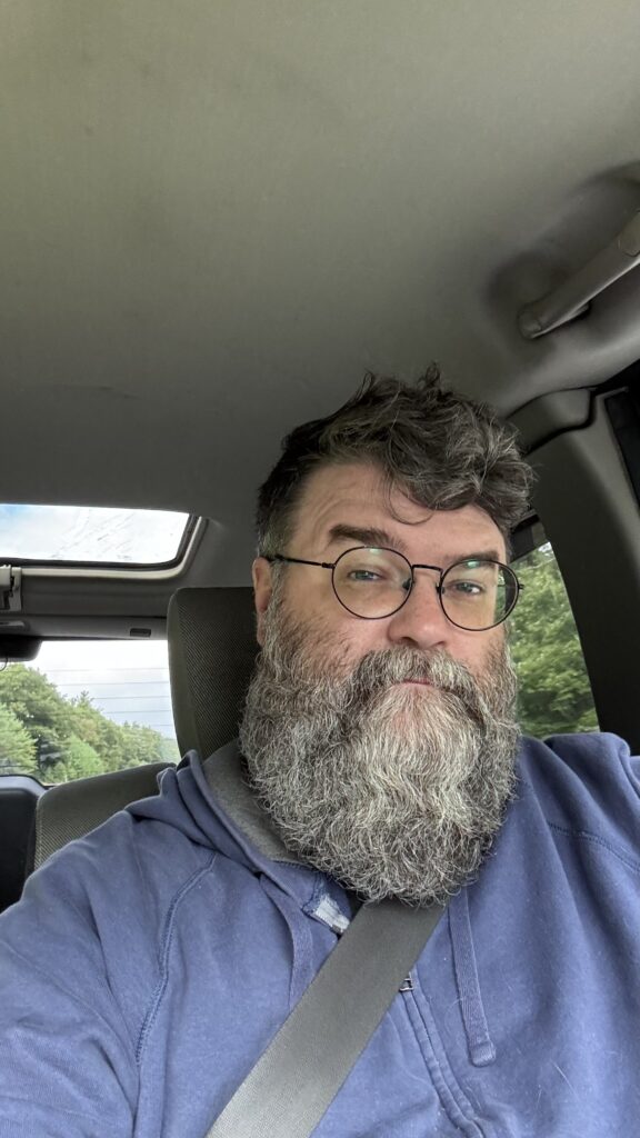 A man with a thick beard and glasses takes a selfie while sitting in the driver's seat of a car. He is wearing a blue hoodie and a seatbelt. The background shows a view of trees and part of a car behind him through the rear window, indicating he is driving on a road surrounded by greenery.