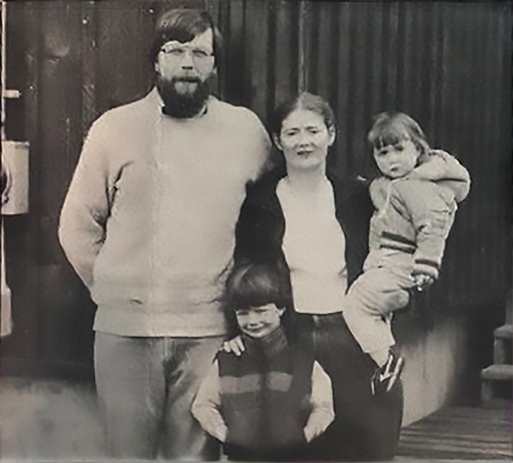 This is a black-and-white photo of a family of four. The father, on the left, has a beard and glasses and wears a light-colored sweater. The mother stands next to him in a dark jacket and white shirt, holding a young child in a winter coat on her hip. In front of the mother is a young boy, Benjamin Jancewicz, smiling slightly and wearing a vest over a long-sleeve shirt. The family stands close together, appearing outdoors near a wooden structure or wall.