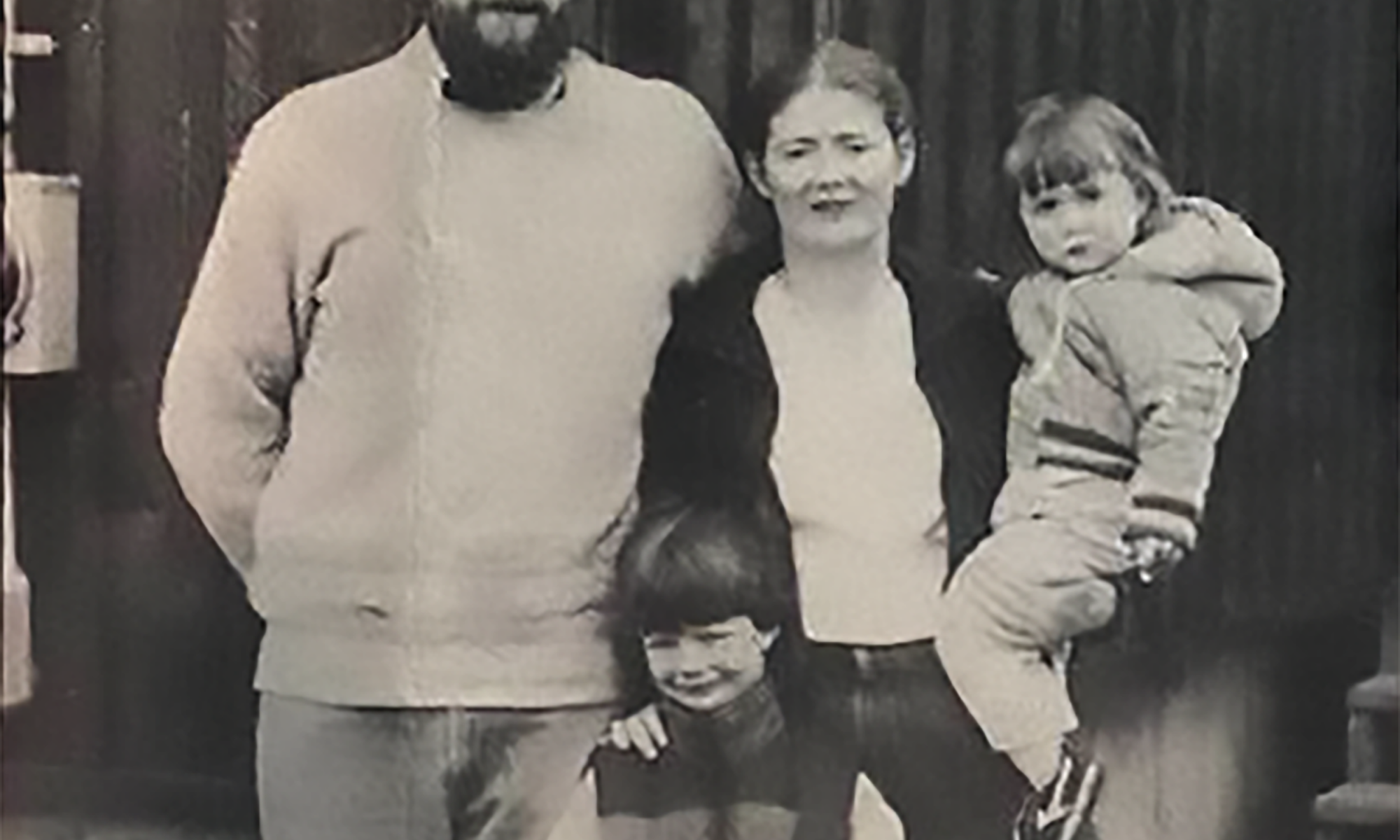 This is a black-and-white photo of a family of four. The father, on the left, has a beard and glasses and wears a light-colored sweater. The mother stands next to him in a dark jacket and white shirt, holding a young child in a winter coat on her hip. In front of the mother is a young boy, Benjamin Jancewicz, smiling slightly and wearing a vest over a long-sleeve shirt. The family stands close together, appearing outdoors near a wooden structure or wall.